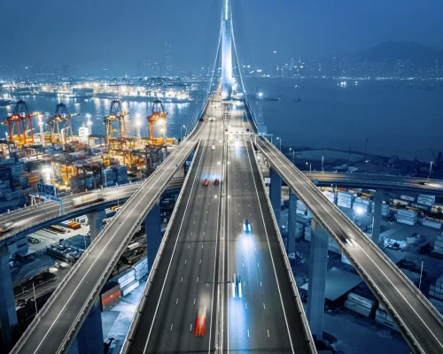 Straße über einem beleuchteten Fracht‑ und Containerhafen bei Nacht.