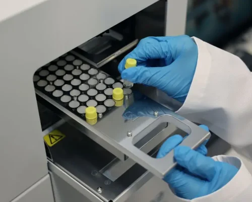 Labortechniker mit blauen Handschuhen setzt eine Probenröhre in ein Analysegerät ein, symbolisch für die Gesundheits‑ und Pharmaindustrie.